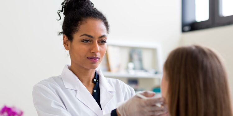 A clinician wearing a white coat and gloves examines a patient’s face during a consultation.